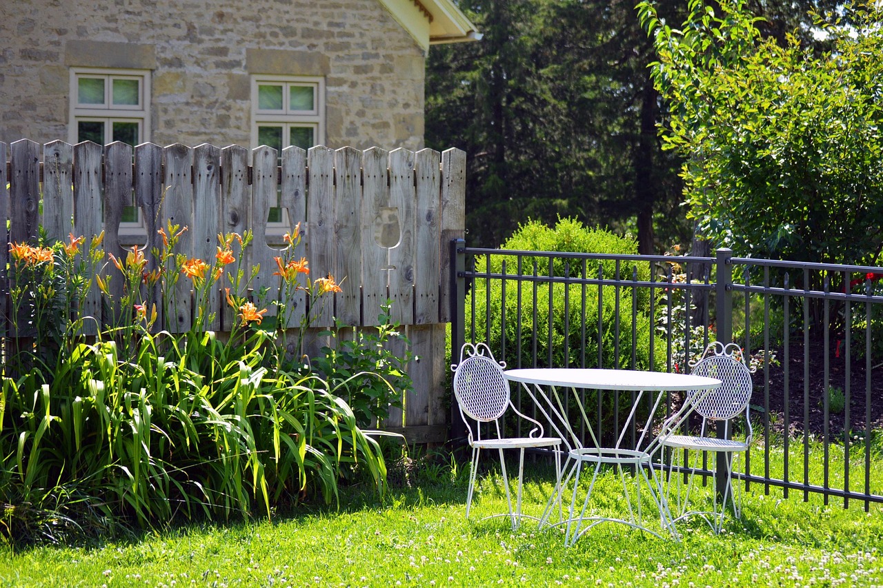 An empty garden bistro set beside a stone house, with daylilies and a wooden fence