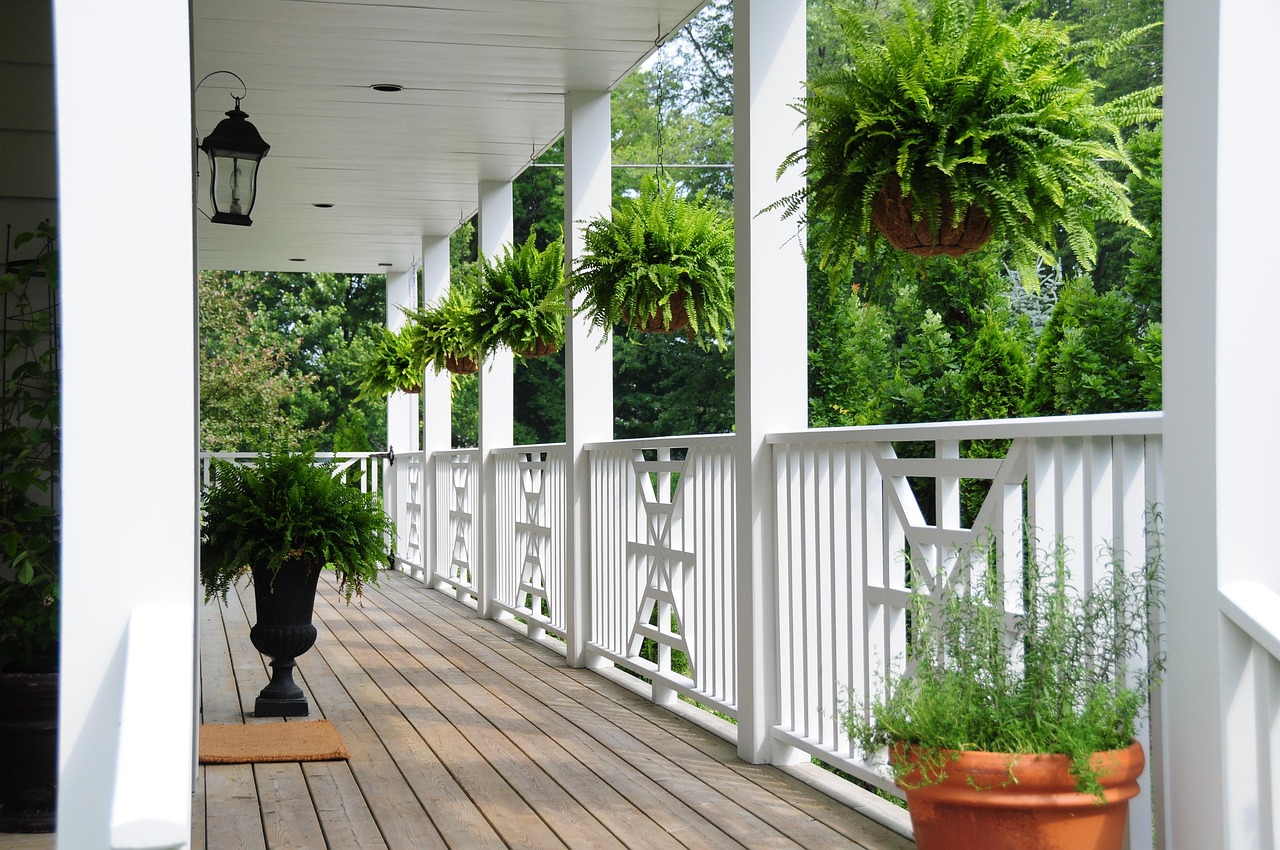 A covered wooden porch with white railings and hanging ferns beside a garden lawn